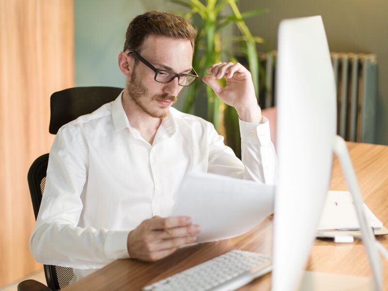 A man seated in front of his computer reads documents with a serious expression, a business owner reading an amended assessment notice from the ATO