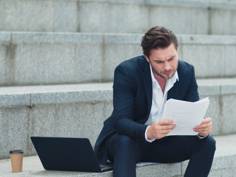 A man in a suit sits on the steps of an outdoor staircase reviewing documents with a stressed expression, a business owner reviewing their general interest charges from the ATO