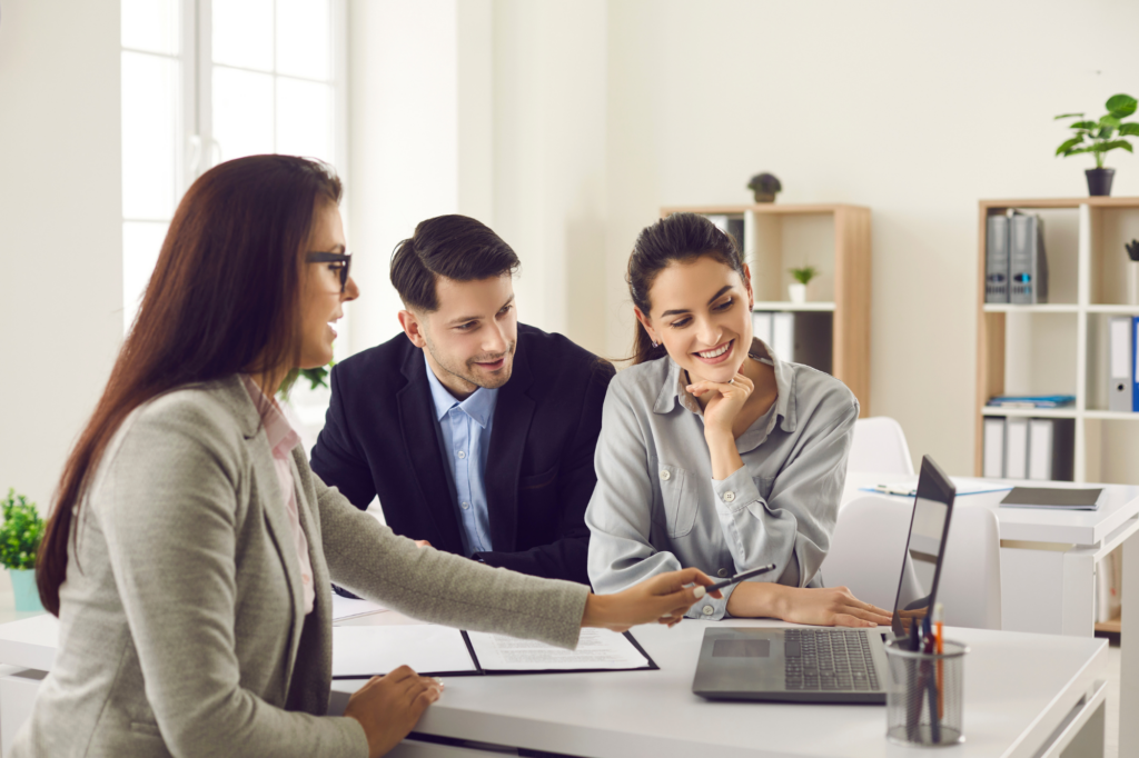 Young business-owning couple being presented a tax debt loan option by a loan expert or loan professional, young couple seated across a woman pointing to the display of her laptop, happy, smiling, solving tax debt problems