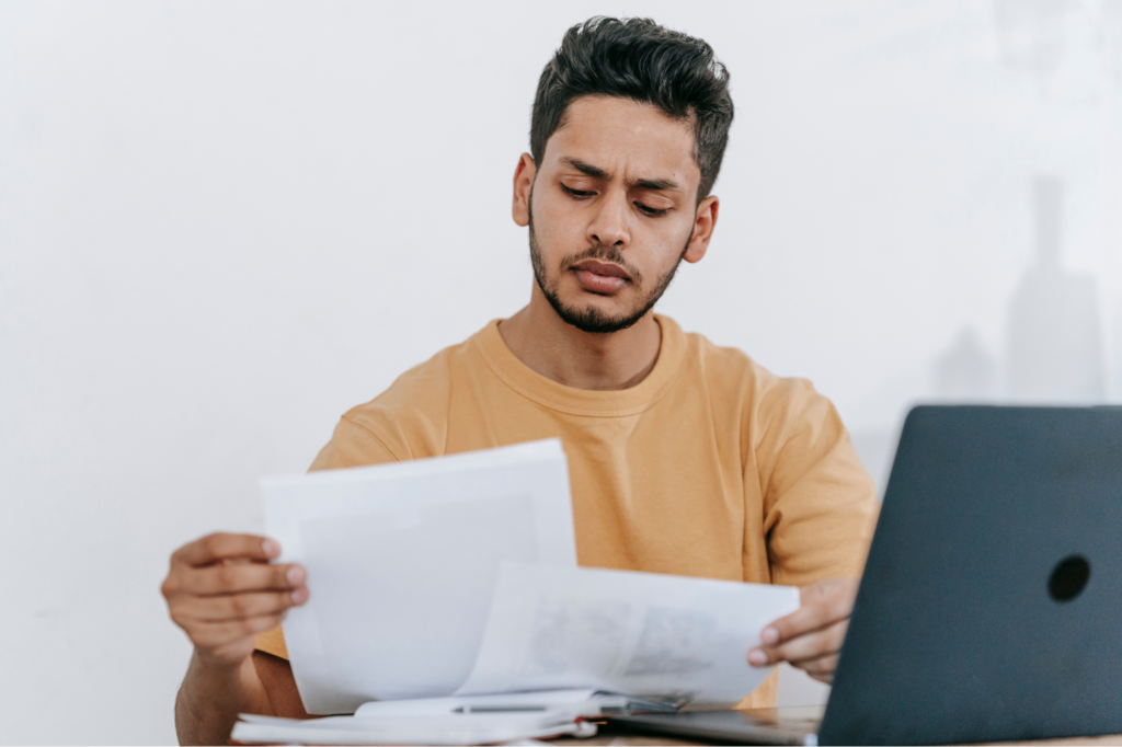 Young adult man concentrating as he reads and compares two documents, sitting in front of a desk with a laptop and a stack of papers, concept photo for considering a loan to pay tax debt