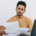 Young adult man concentrating as he reads and compares two documents, sitting in front of a desk with a laptop and a stack of papers, concept photo for considering a loan to pay tax debt