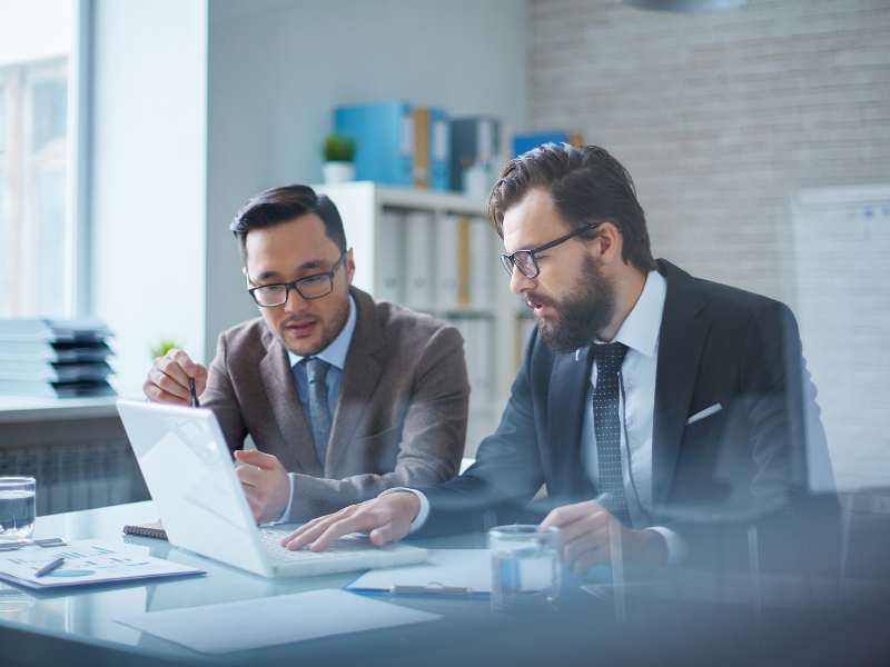 Two business partners intently discussing a buyout, two men in suits and glasses sit in a professional setting and discuss what’s on the laptop