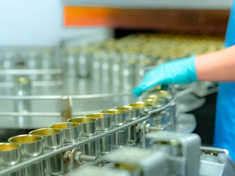 A factory worker’s hand guides a line of cans going through a large factory machine, canned food factory