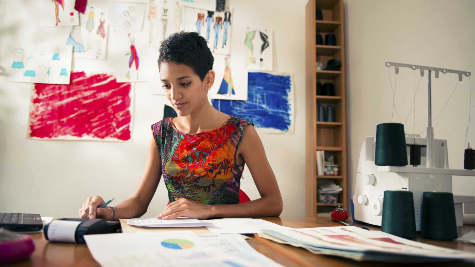 Woman with a fashion business, sitting down working on business finance calculations, fashion images and sewing materials visible