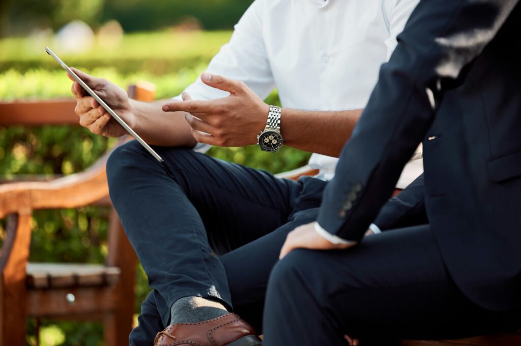 Outdoor setting, cropped photo of two men in business attire sitting on a bench, discussing business finance using an electronic tablet