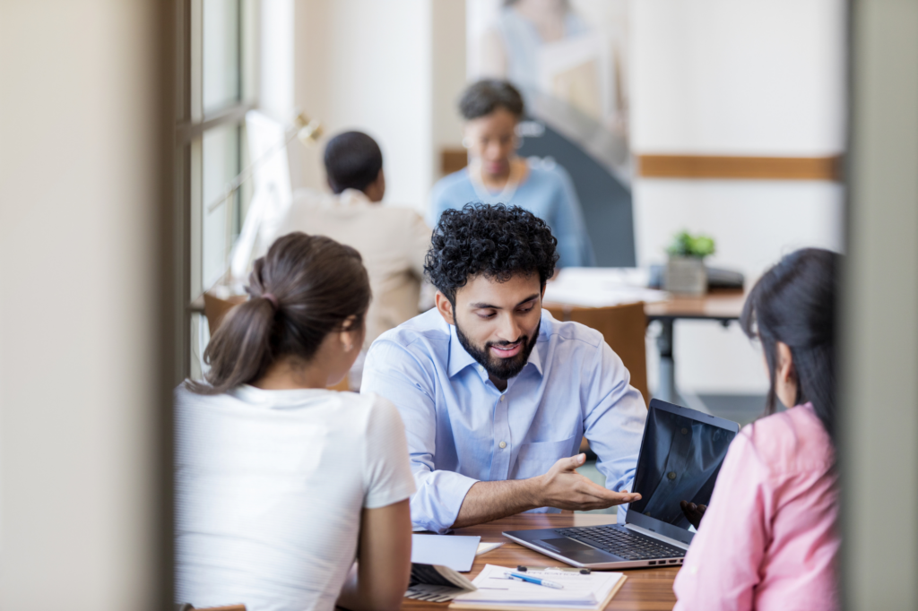 Two women sit across a man discussing what’s on the display of his laptop, concept photo for a lender or loan expert discussing unsecured business loan details with business owners