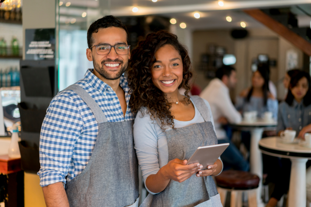 A man and woman wearing casual clothes and aprons smile at the camera. The business-owning couple is happy posing inside their cafe business, with the woman holding a tablet. Concept photo for getting a no-doc or low-doc loan approved for business.