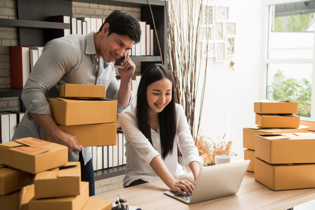 A man and woman smile while reading the laptop monitor. The woman is sitting down with her fingers on the keyboard, the man is standing, holding boxes in one hand and a phone in the other. Concept photo for getting a no-doc or low-doc loan for a small business.