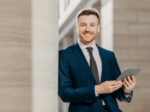 A business owner in a blue suit smiling at the camera, leaning against the wall and holding an electronic tablet