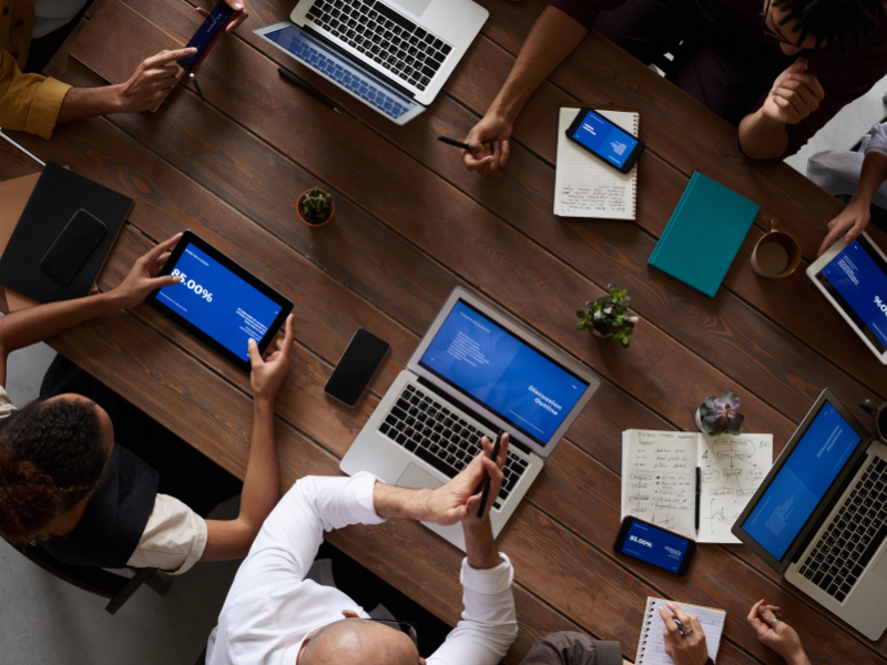 Top view of a conference table with various business stakeholders having a meeting with their phones, tablets, and laptops