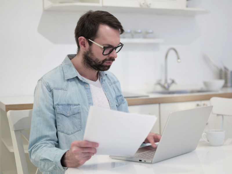 A man in glasses uses his laptop while holding documents in one hand