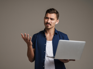 A man with a puzzled expression and a hand thrown up in question, other hand holding a laptop, grey background