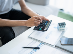 Cropped photo of a man using a calculator, documents, a notebook, a phone, and a miniature house on the table, borrower calculating the cost of a second mortgage loan