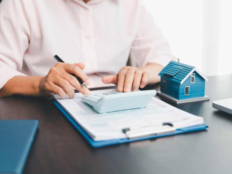 Cropped photo of a man using a calculator, a clipboard of files and a miniature house visible on the table