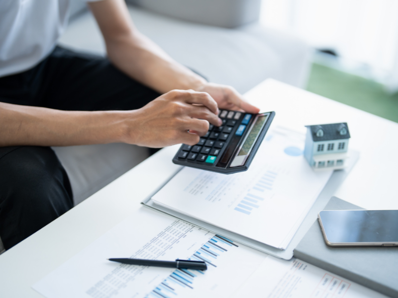 A man using a calculator, scattered documents, a pen, phone, notebook, and a miniature house on the table