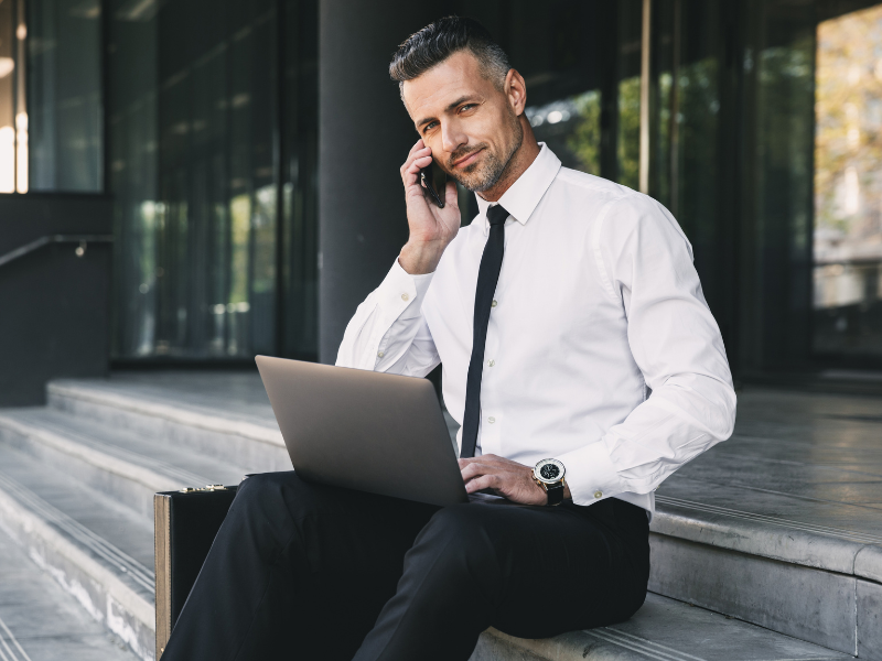 A business owner sitting on the stairs outside of a building, talking on the phone with his laptop on his lap, looking at the camera