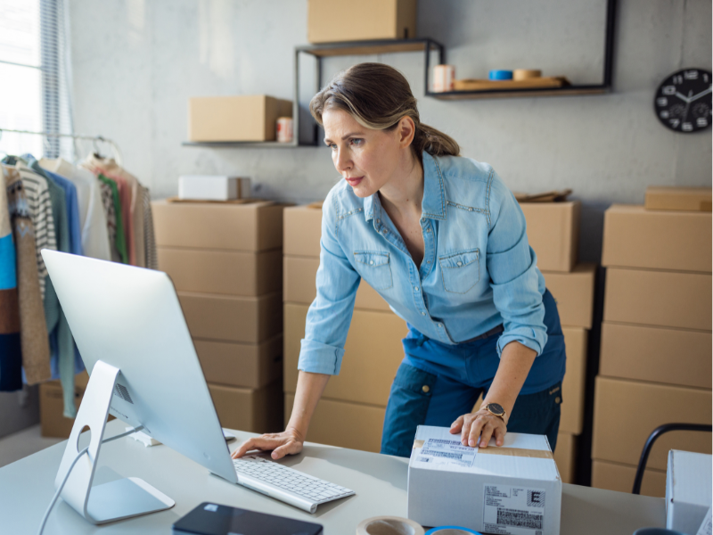 Business owner works on her computer while standing up, one hand on top of a delivery box