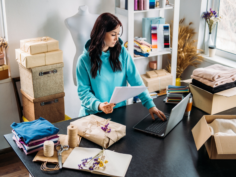 A small business owner reading documents and working on her laptop, surrounded by products and packaging materials