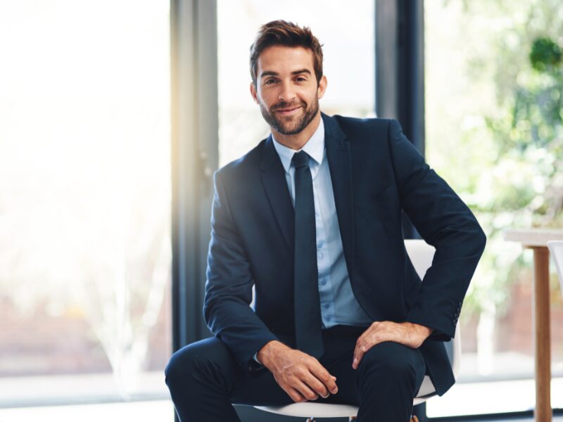 A man in a suit sitting on a chair posing for a photo with one hand on his thigh, smiling business owner