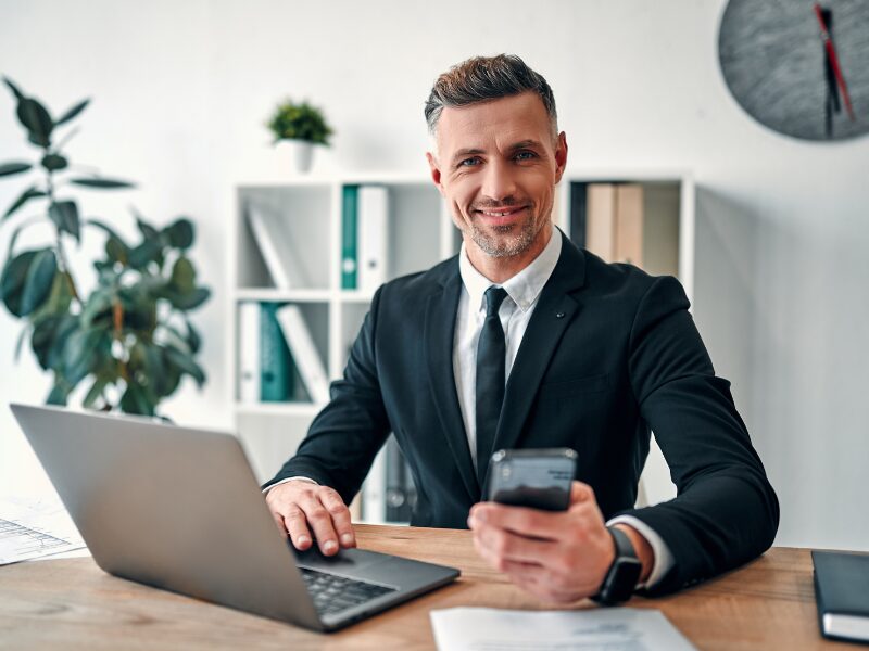 A professional man in a suit looks directly at the camera while holding his smartphone and using his laptop, a business owner happy about his finances