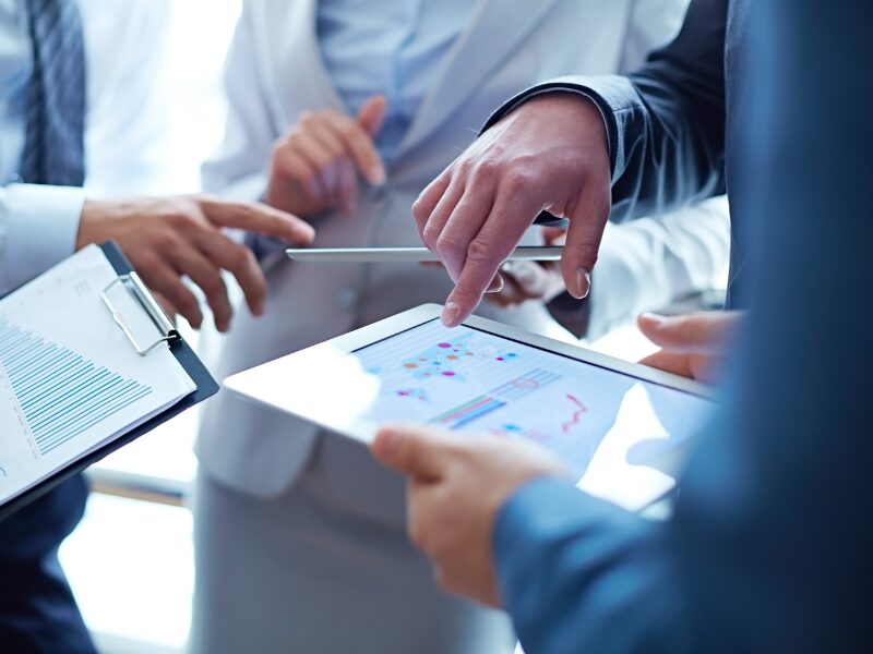 People working in a business discuss growth strategies while holding electronic tablets and clipboards with documents