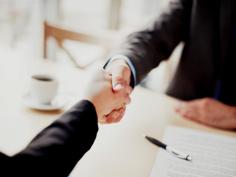 Two men shaking hands, a cup of coffee, a document, and a pen on the table below