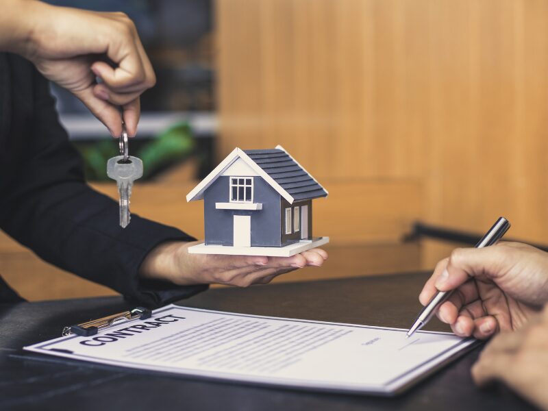 A mortgage broker or lender holds up a home key in one hand and a miniature house in the other while the client signs the loan contract