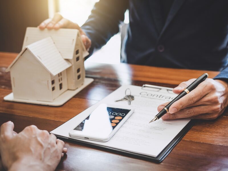 A loan broker or lender holds a pen in one hand and a miniature house in the other, presenting a loan contract, house keys, and a smartphone displaying the calculator app to the borrower