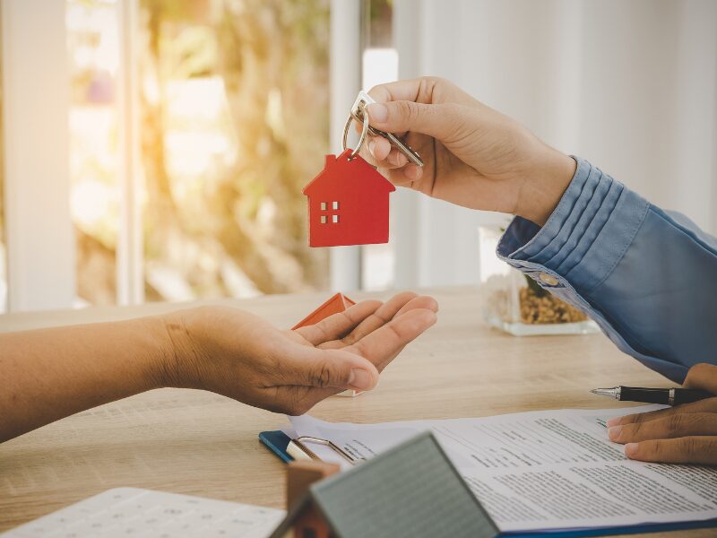 A loan broker or lender hands a house key with a house-shaped red keychain to a client after signing the loan agreement