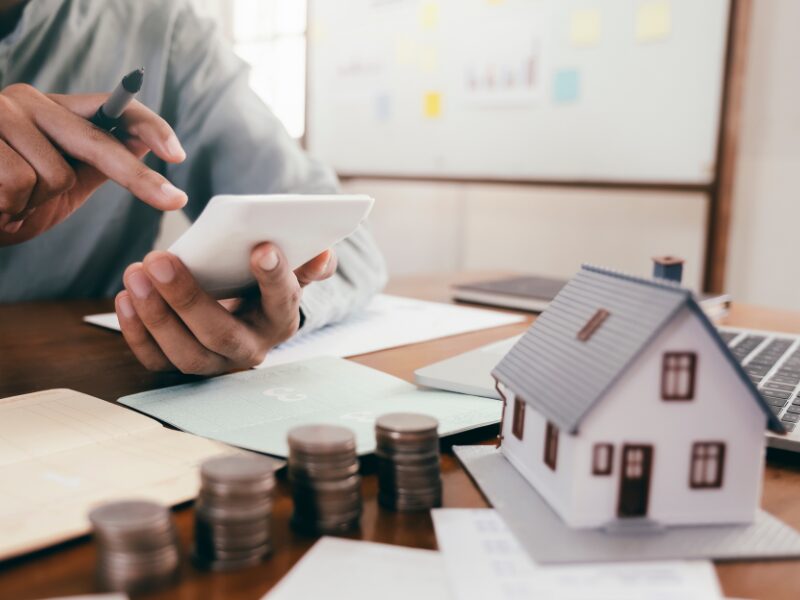 A borrower uses a calculator to see the full cost of a loan, bankbooks, coins, a laptop, documents, a notebook, and a miniature model house on the table