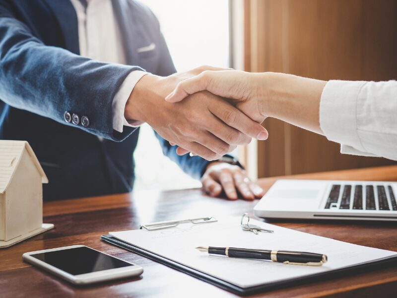 A mortgage broker or a lender shakes hands with a borrower after they sign the loan agreement, a miniature house, smartphone, loan contract, fountain pen, laptop, and house keys on the table below