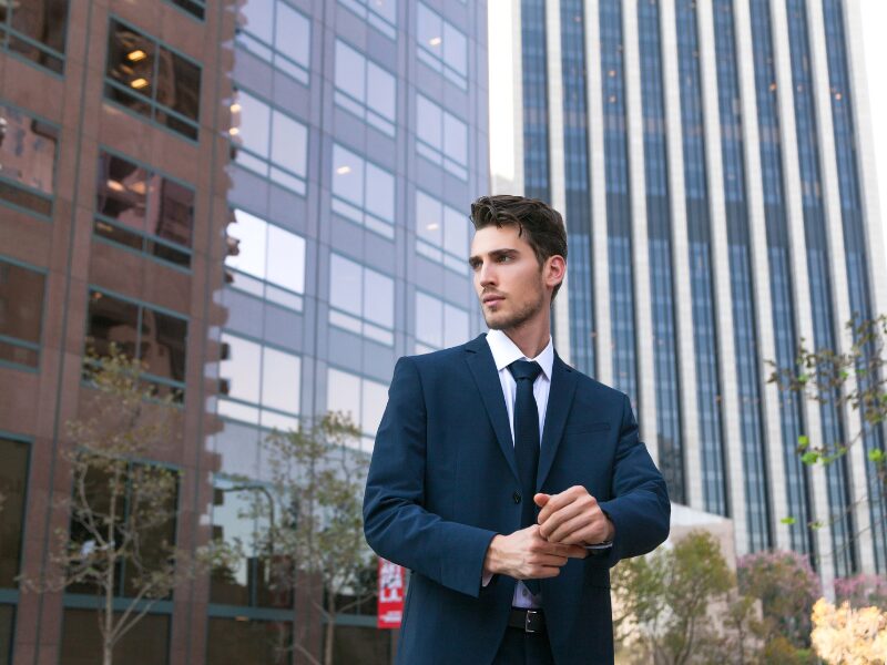 A businessman in a suit adjusts his shirt cuff, standing outdoors surrounded by buildings