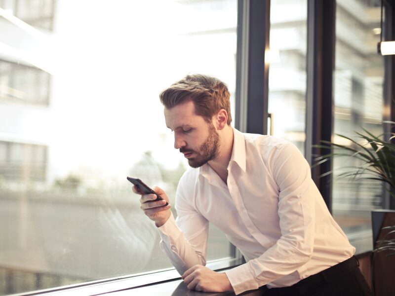 A man in a professional setting leans by the window while reading news updates on his smartphone