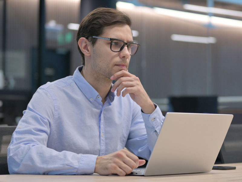 A man in glasses with his hand on his chin, thinking while using his laptop, choosing between a secured and an unsecured loan