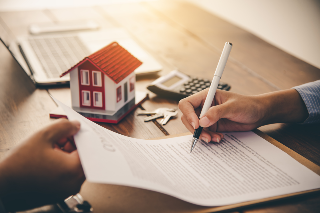 Cropped photo, loan broker handing refinancing documents to client, client signing, table with documents, calculator, and miniature model house, concept photo