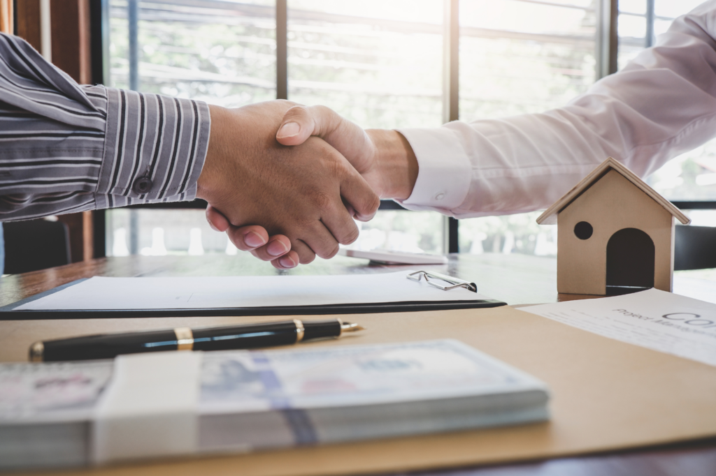 Cropped photo of a handshake, client and mortgage broker agree on SMSF loan services, files, documents, and wooden model house visible on desk, concept photo