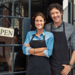 Couple posing happily at the store front of their small business, open sign hanging up