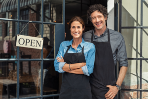Couple posing happily at the store front of their small business, open sign hanging up
