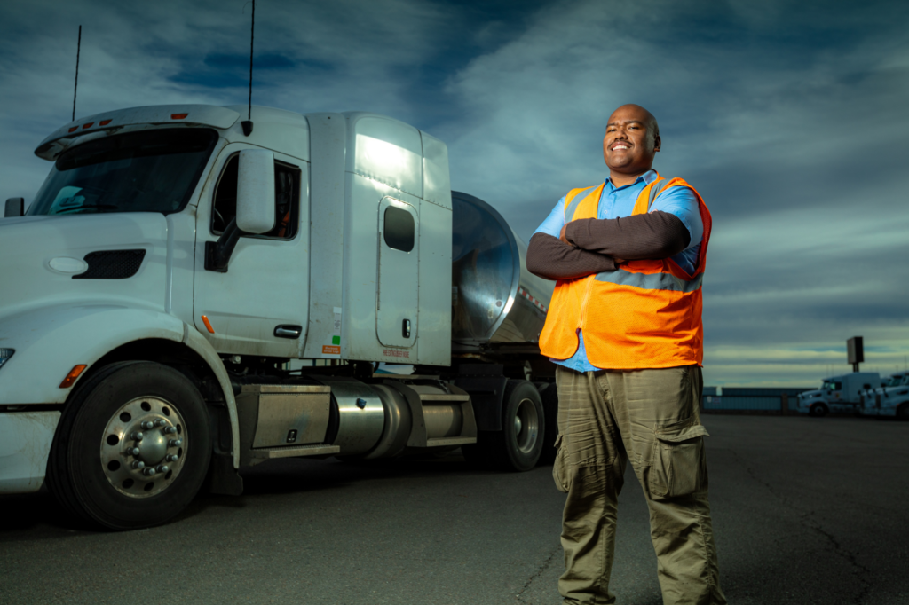 Large and cheerful truck driver or business owner poses in front of his truck, smiling man in blue shirt and hi-visibility vest, concept photo for asset based commercial loans using trucks