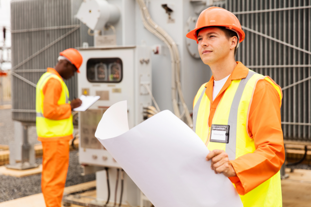 Data and electrical engineers in full high visibility attire and hard hat take checks at a site, one man checking wire box, one man holding plans while looking hopefully into the distance