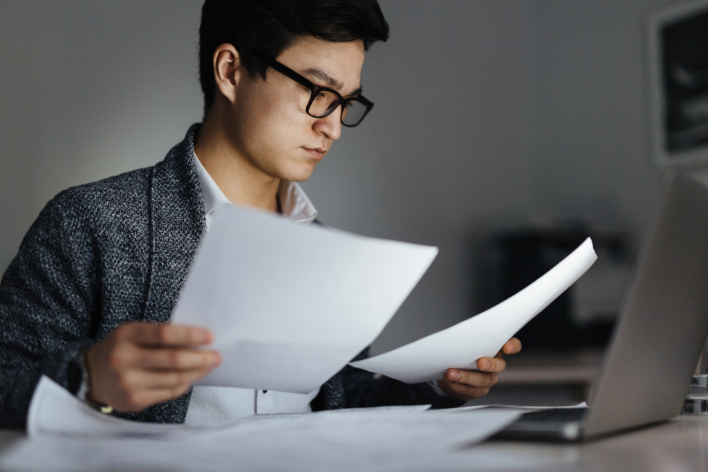 Young professional in dark-rimmed glasses chooses between two documents, concentrating, seated in front of an open laptop, concept photo of a business owner choosing between business debt consolidation vs bankruptcy