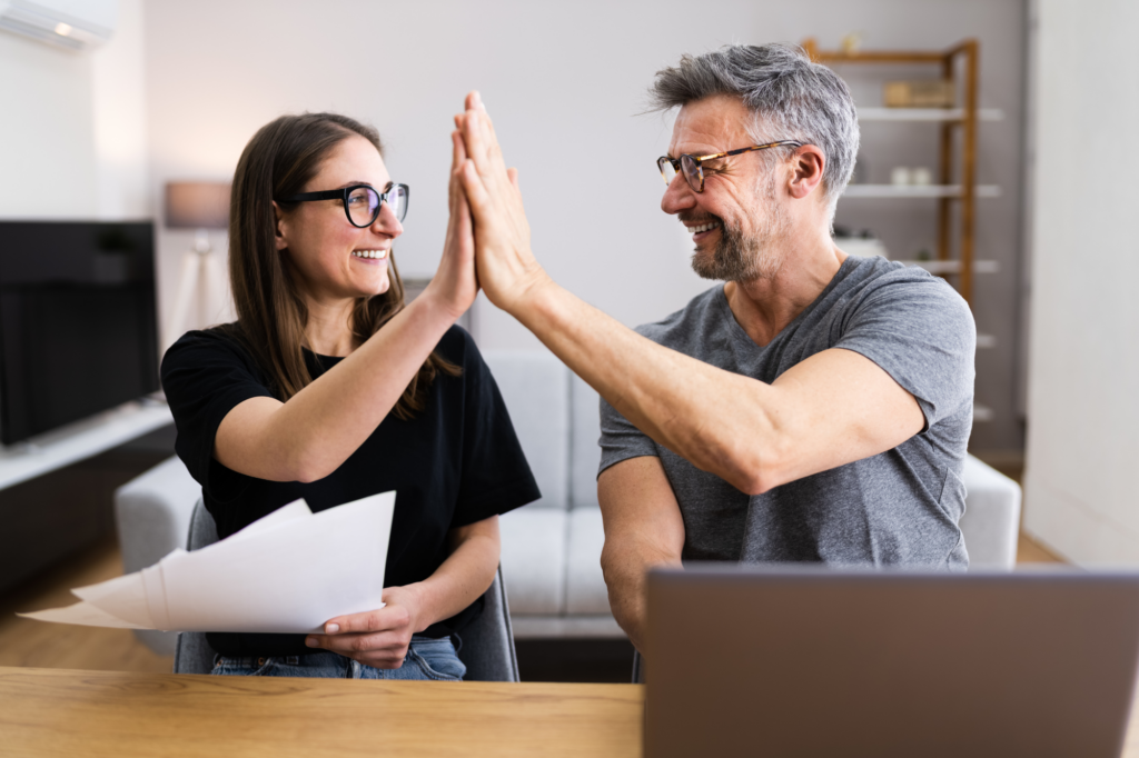 Couple high–fives, smiling at each other, the woman is holding a few pieces of paper, concept photo for business partners or business owners celebrating after their business loan application is approved