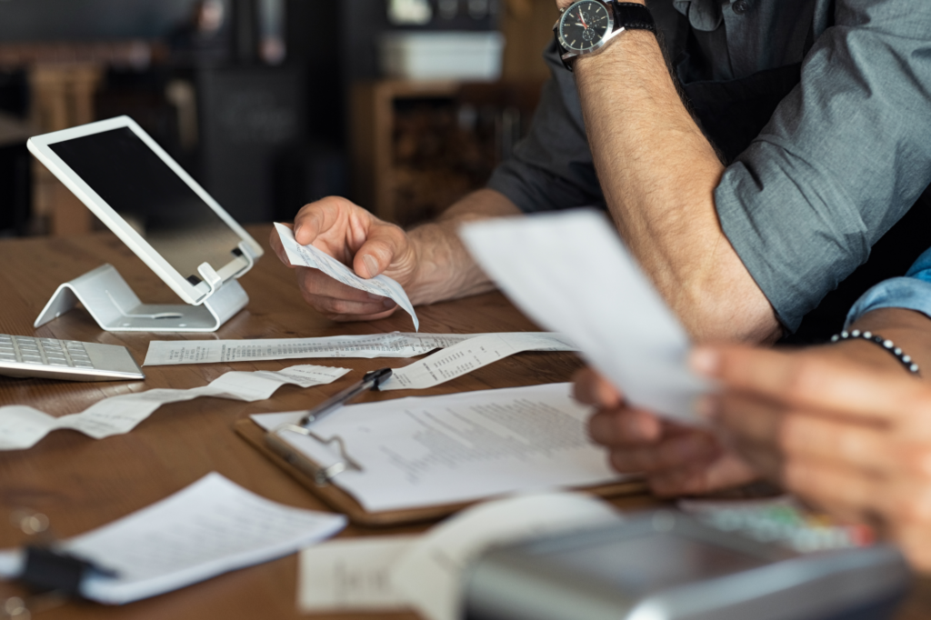 Cropped side view photo of business partners examining tax documents, invoices, financials, and other documents, papers and tablet visible on wooden table