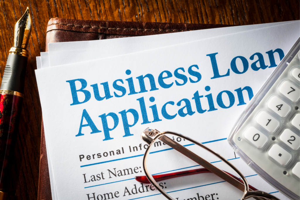 Zoomed and cropped photo of an assortment of objects, the focus is on the corner of a document saying Business Loan Application in big, blue letters in a serif font, with a leather folder, reading glasses, a calculator, and a fountain pen arranged around the paper