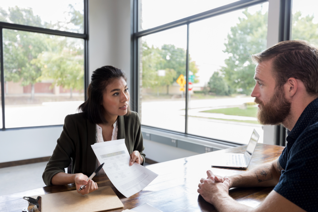 A loan broker or lender shows a business owner the requirements and details for a business loan in Australia, woman in business attire uses pen to point on a piece of paper she’s showing to a man sitting across the table