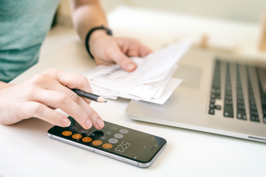 Cropped photo of a man calculating household expenses, holding recepits, typing into calculator app in phone, laptop visible on table