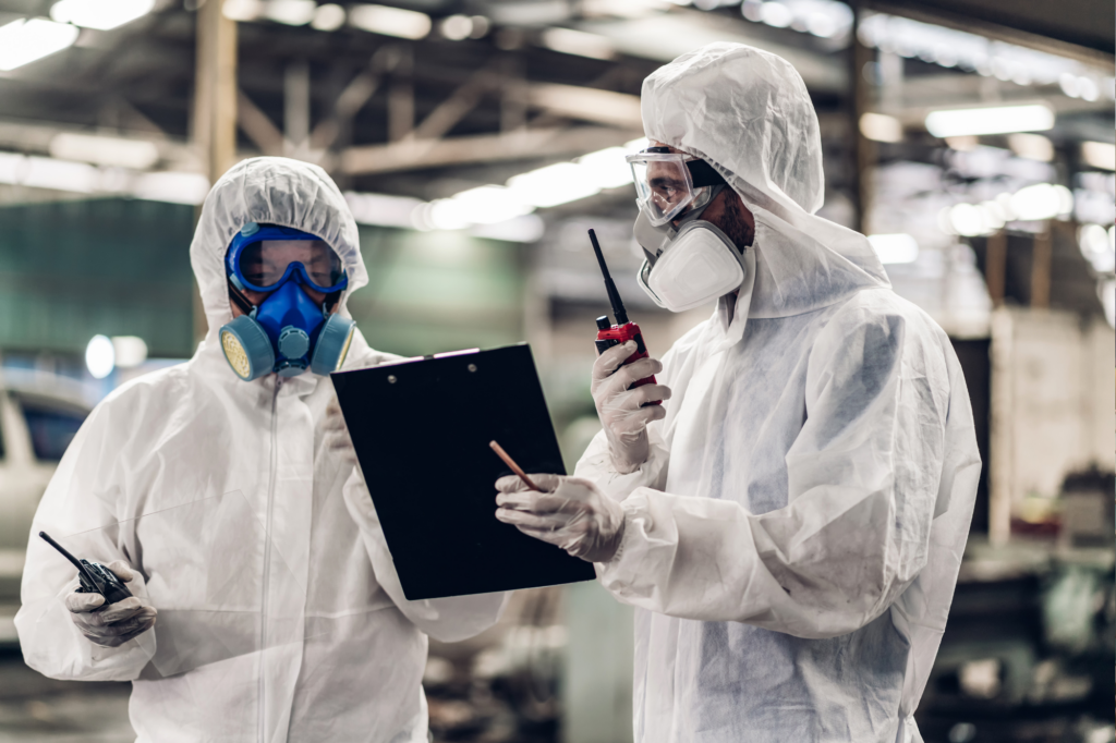 Two chemicals manufacturing factory workers converse while wearing full PPE and looking at a clipboard. One of the workers has a comunications radio in his hand