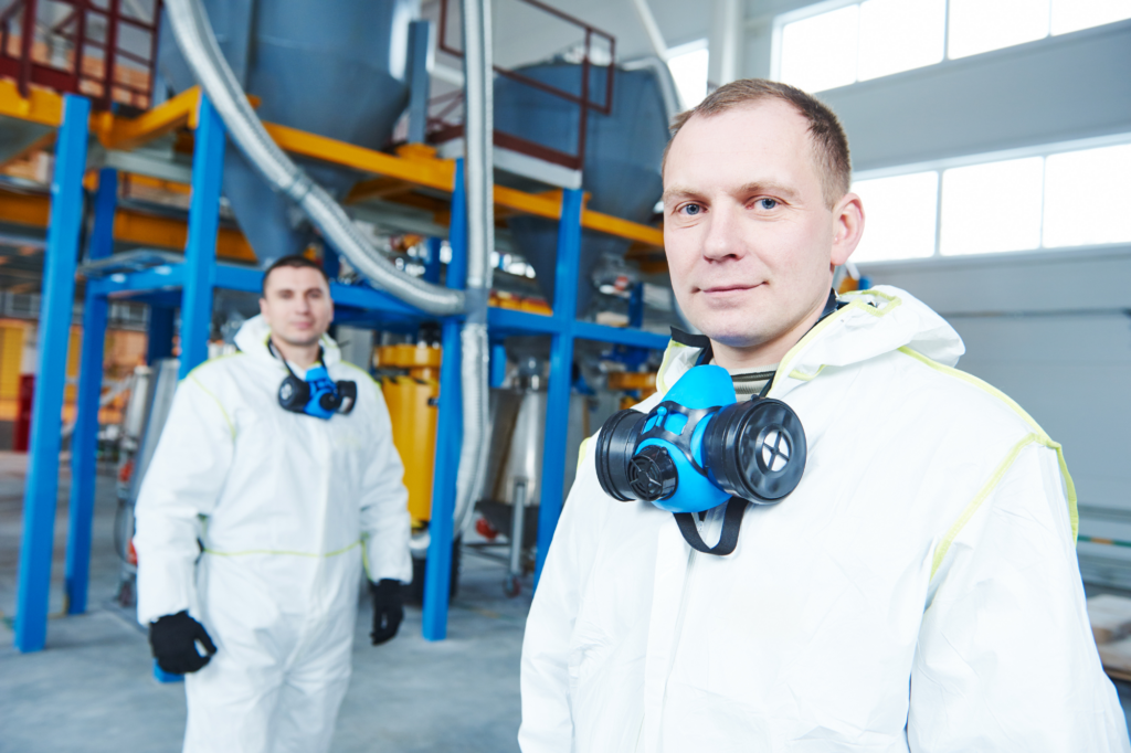 Workers in a chemicals manufacturing plant wearing PPE and gas masks pulled down, looking at camera and smiling, concept photo for chemicals manufacturer getting a loan structure approved