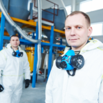 Workers in a chemicals manufacturing plant wearing PPE and gas masks pulled down, looking at camera and smiling, concept photo for chemicals manufacturer getting a loan structure approved