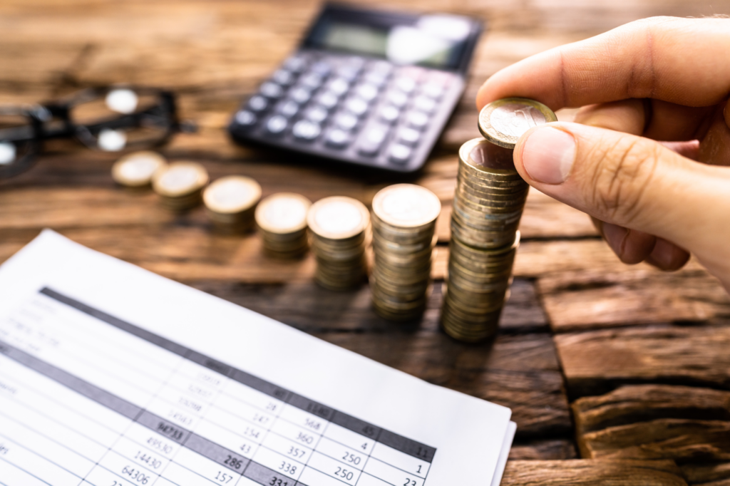 Zoomed in photo of a hand stacking up coins on a wooden table, with documents, calculator, and glasses also visible, concept photo of using personal resources or savings for business